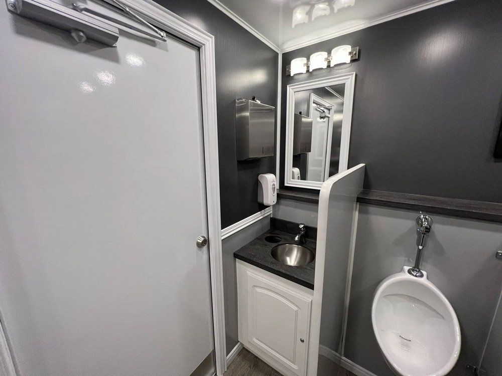 Interior of a luxury portable restroom with sink, urinal, and mirror; gray and white color scheme.