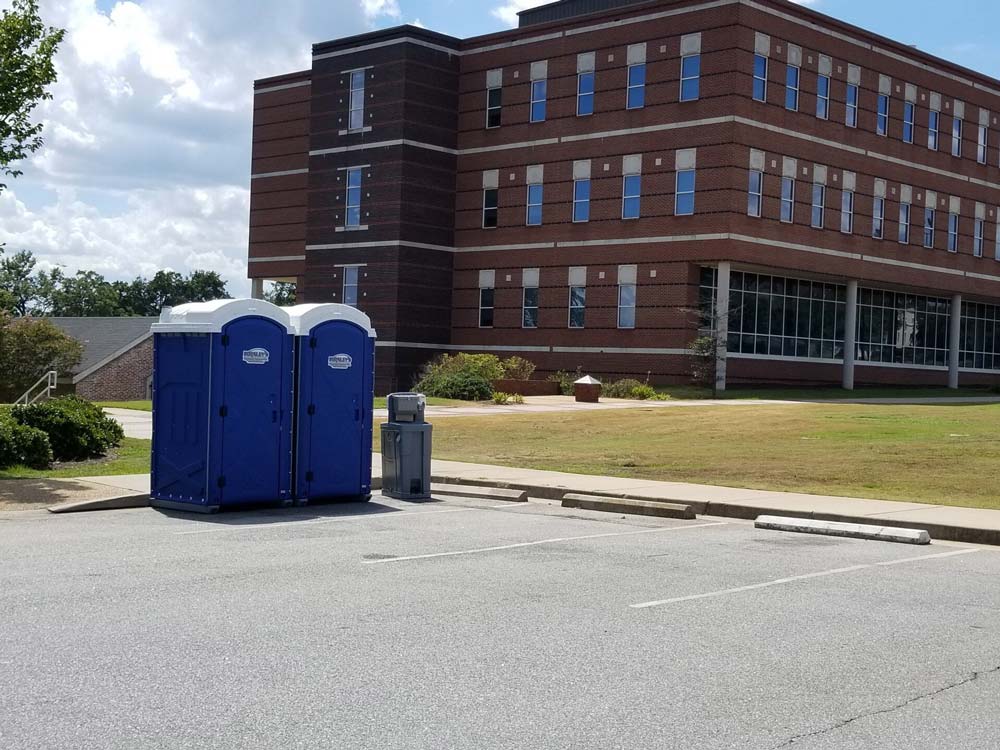 Two blue portable toilets in a parking lot in front of a brick building.