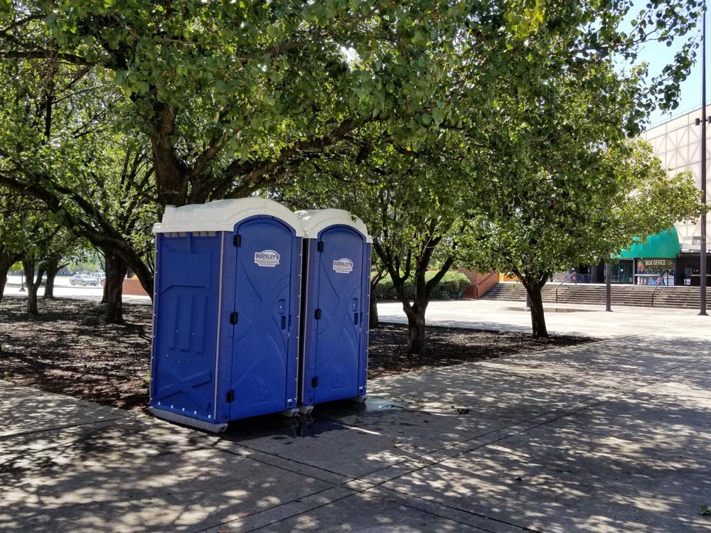 Two blue portable toilets under trees on a paved area.