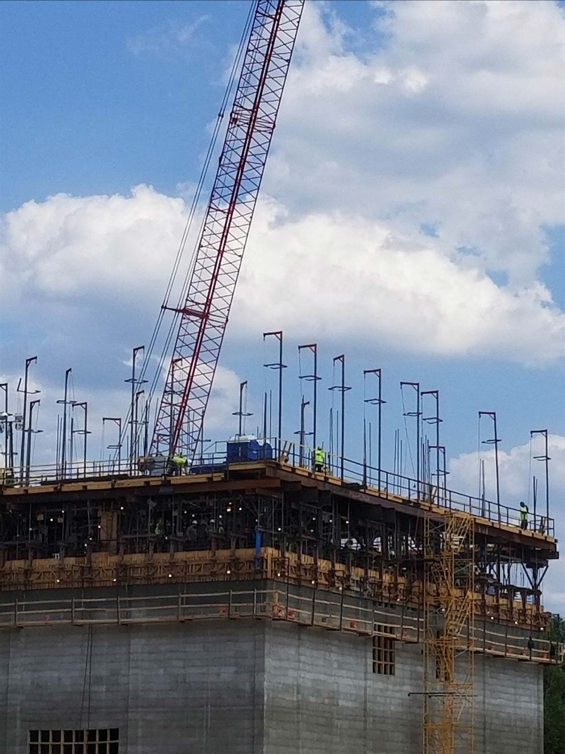 Construction site with crane reaching a building's upper level; workers present; blue sky and clouds in the background.