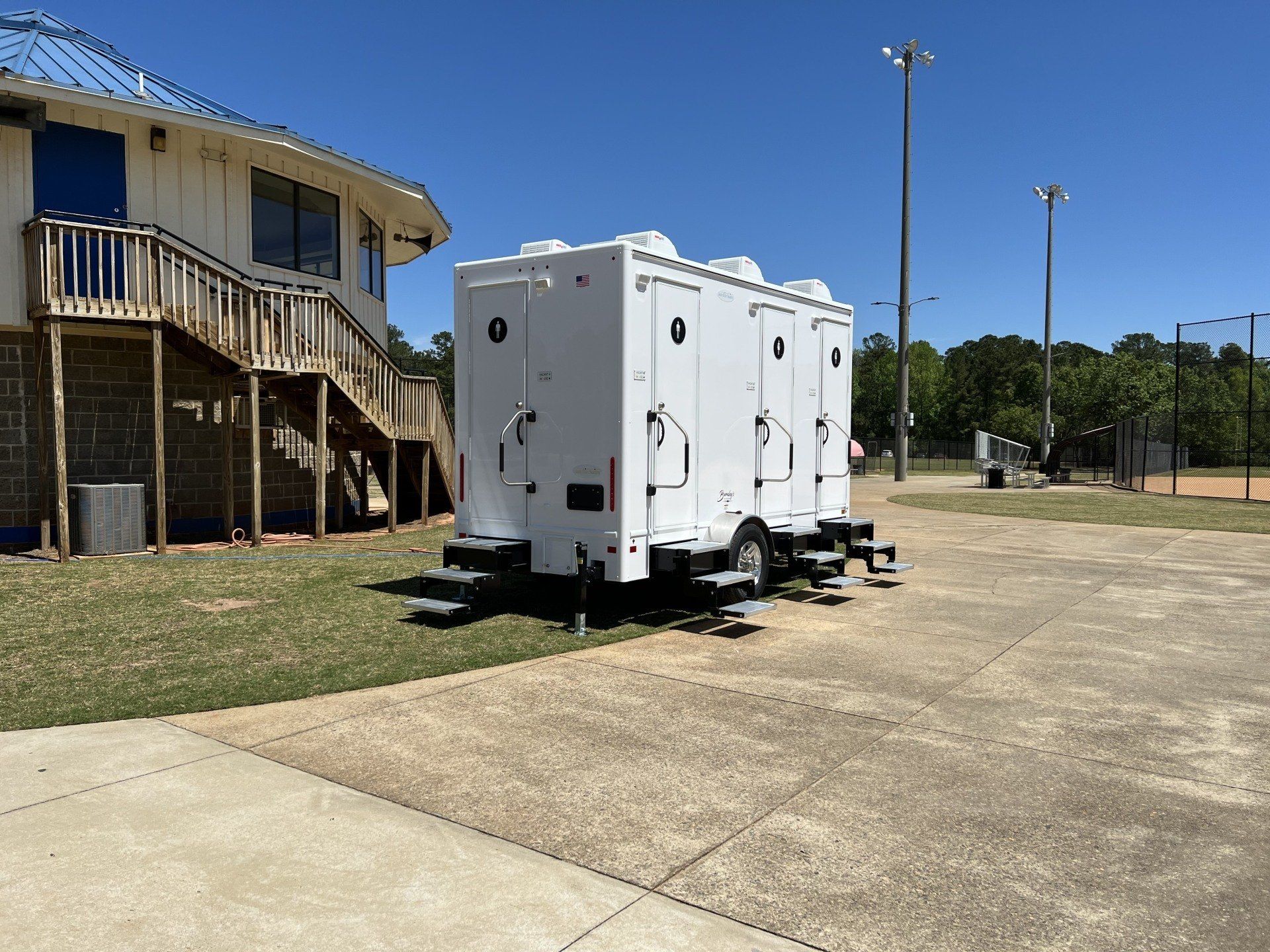 White portable restroom trailer on concrete, next to a building with steps, under a blue sky.