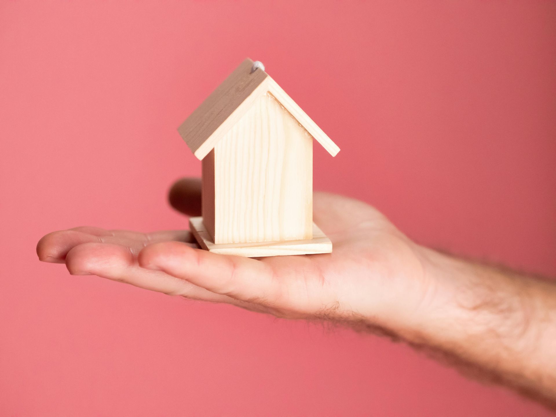 Hand holding a small wooden house against a pink background.
