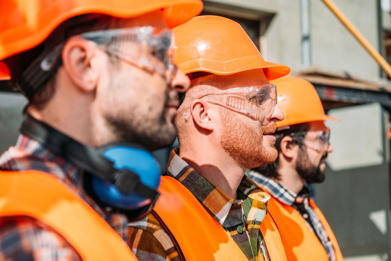 Tre operai edili con elmetti arancioni e occhiali di sicurezza in un cantiere, visti di profilo.