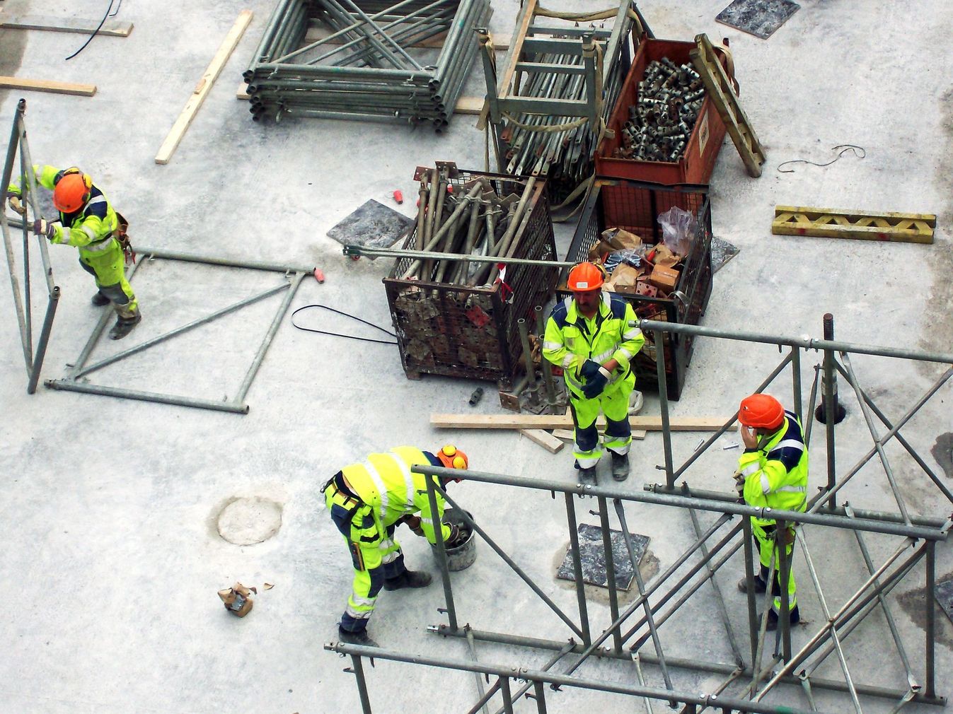Operai edili con caschi arancioni assemblano impalcature metalliche in un cantiere di cemento.