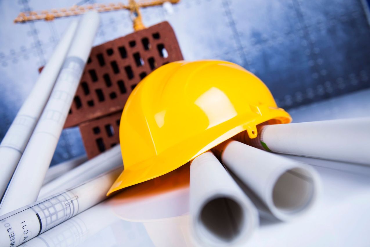 Casco giallo da cantiere appoggiato su progetti e tubi da costruzione arrotolati, con mattoni sullo sfondo.