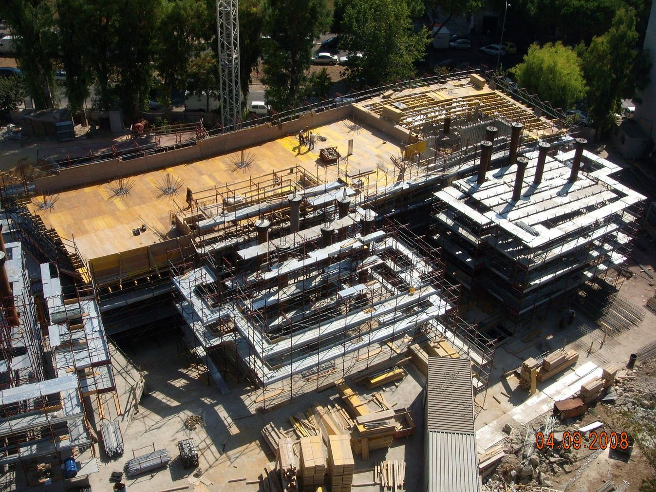 Veduta aerea di un grande cantiere edile con struttura in acciaio, pavimentazione in legno e veicoli parcheggiati.