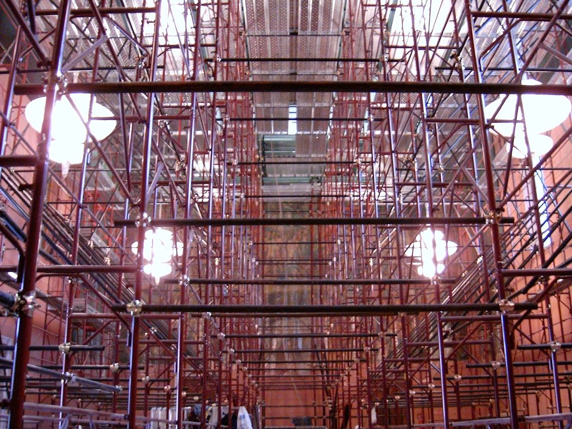 Ponteggi e struttura metallica all'interno di un grande cantiere al coperto con luci intense.