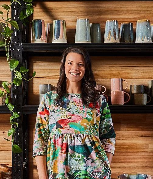 A woman in a colorful dress is standing in front of a shelf full of mugs.