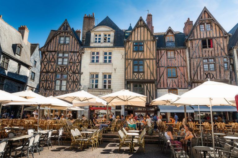 A group of people are sitting at tables under umbrellas in front of a row of buildings.