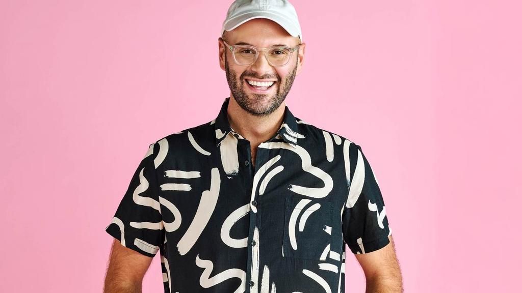 A man wearing a hat and glasses is standing in front of a pink background.