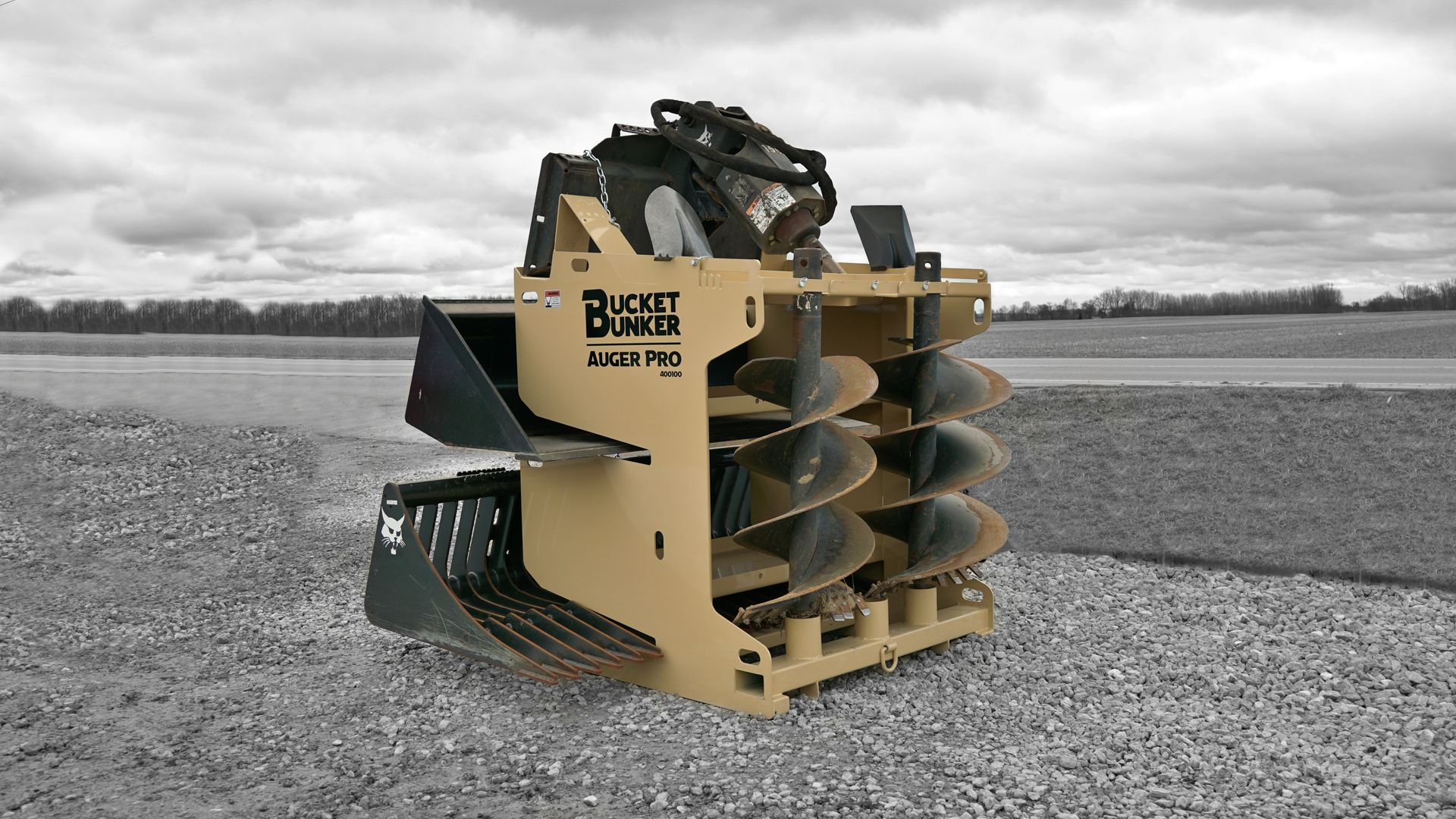 A bucket bunker with a buckets, auger bits, and auger drive attached to it is sitting on a gravel road.