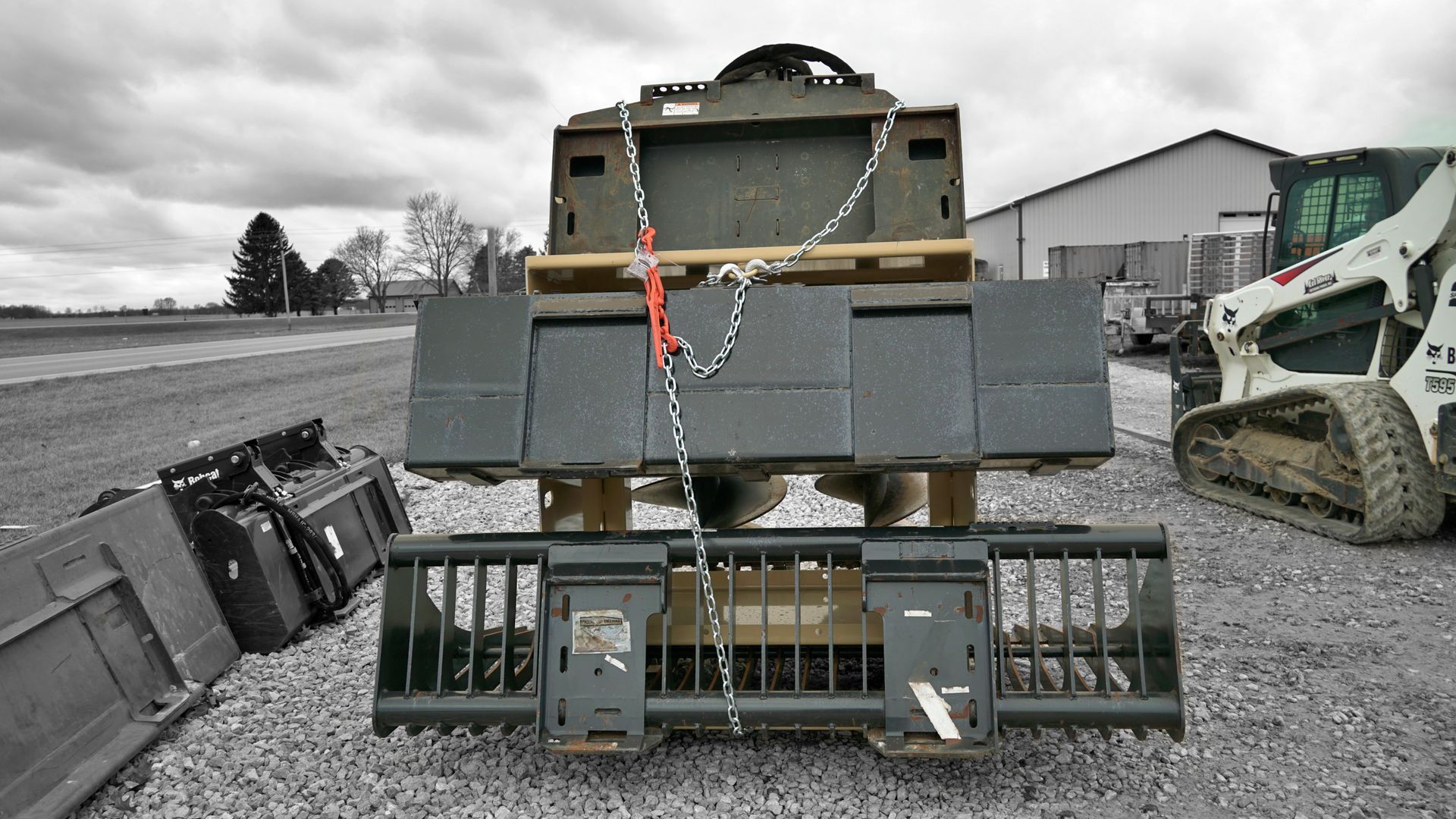A tractor is parked in a gravel lot next to a bucket bunker attachment rack loaded with attachments. 