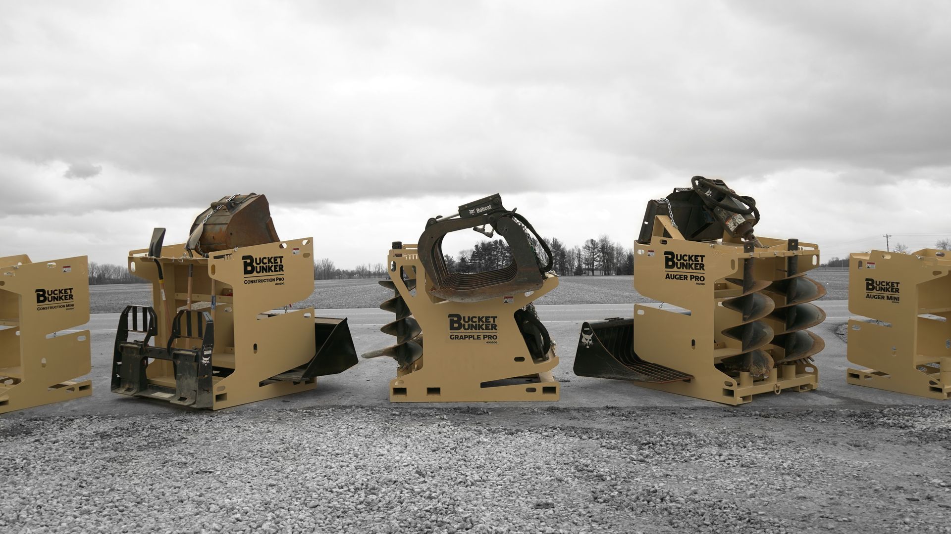 Three bucket bunkers sitting in a gravel lot with attachments on them like grapples, auger drive, auger bits, etc. 