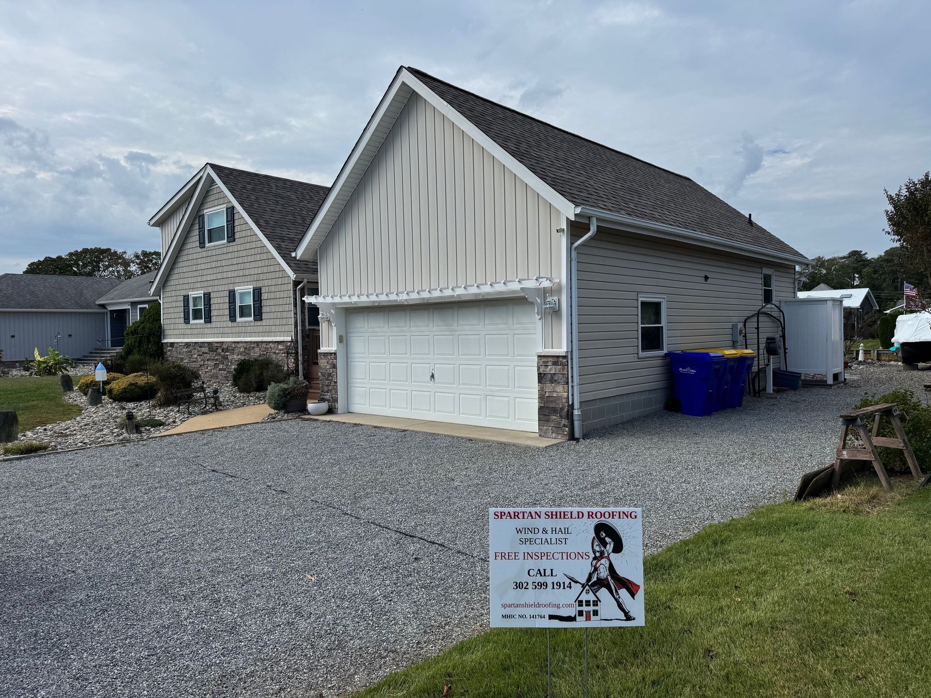 Gravel driveway with a gray garage and house, blue trash bins, and a yard sign under a cloudy sky