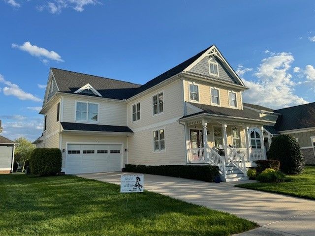 Two-story cream house with white trim, front porch, driveway, and a green lawn under a blue sky.