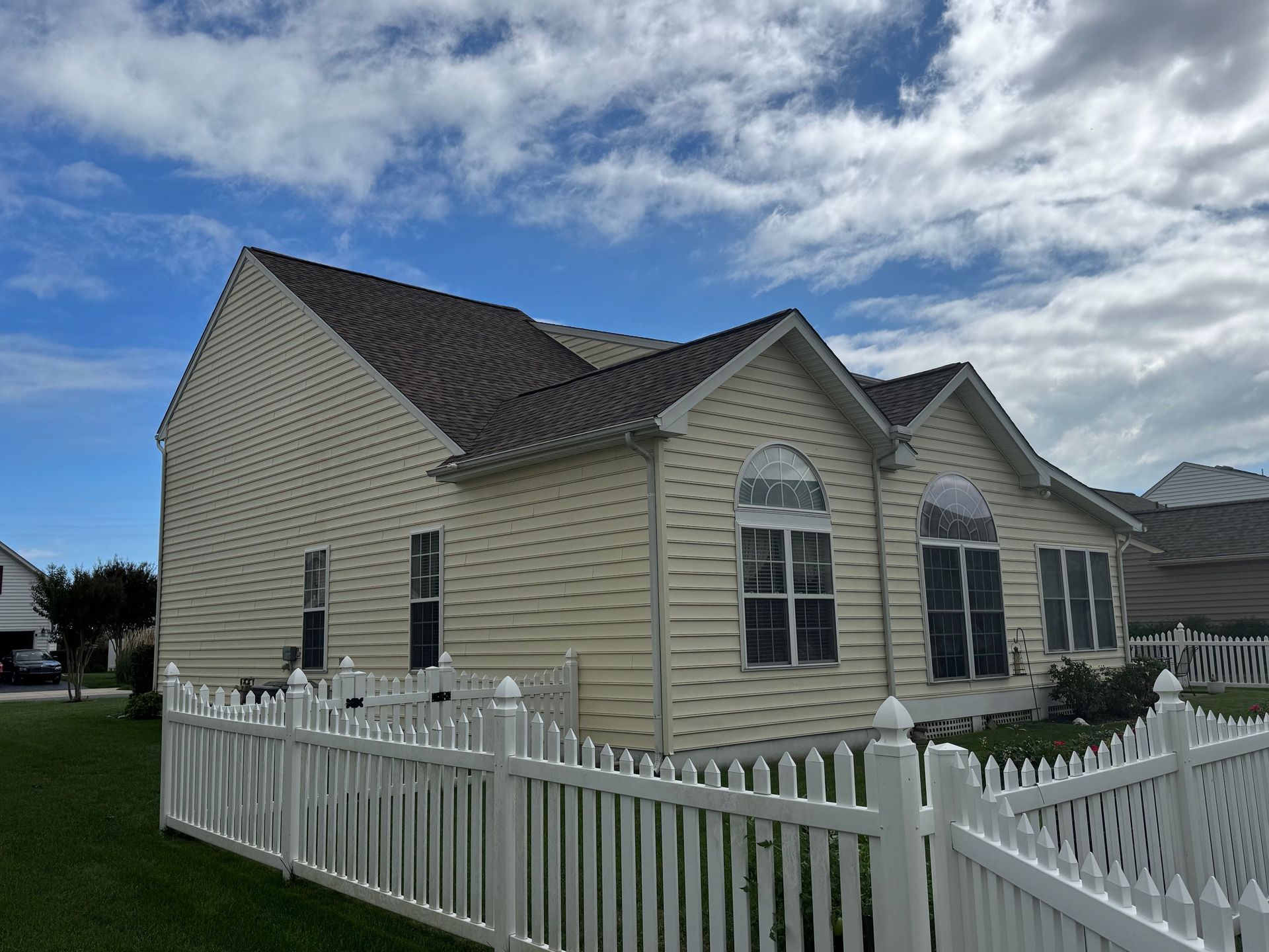 Beige house with dark roof and white picket fence under a partly cloudy blue sky