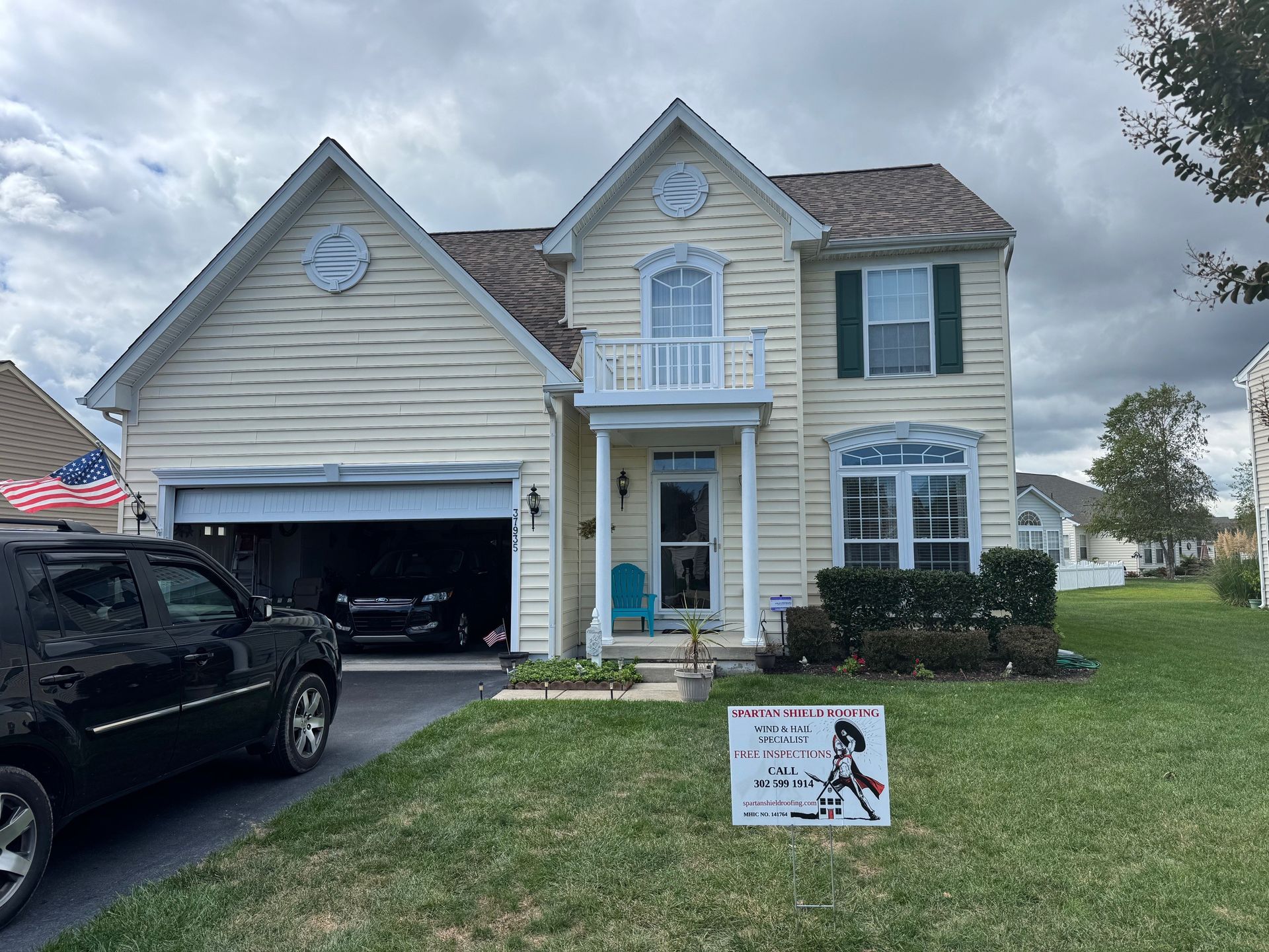 Two-story beige house with an open garage, parked SUV, and a lawn sign under a cloudy sky