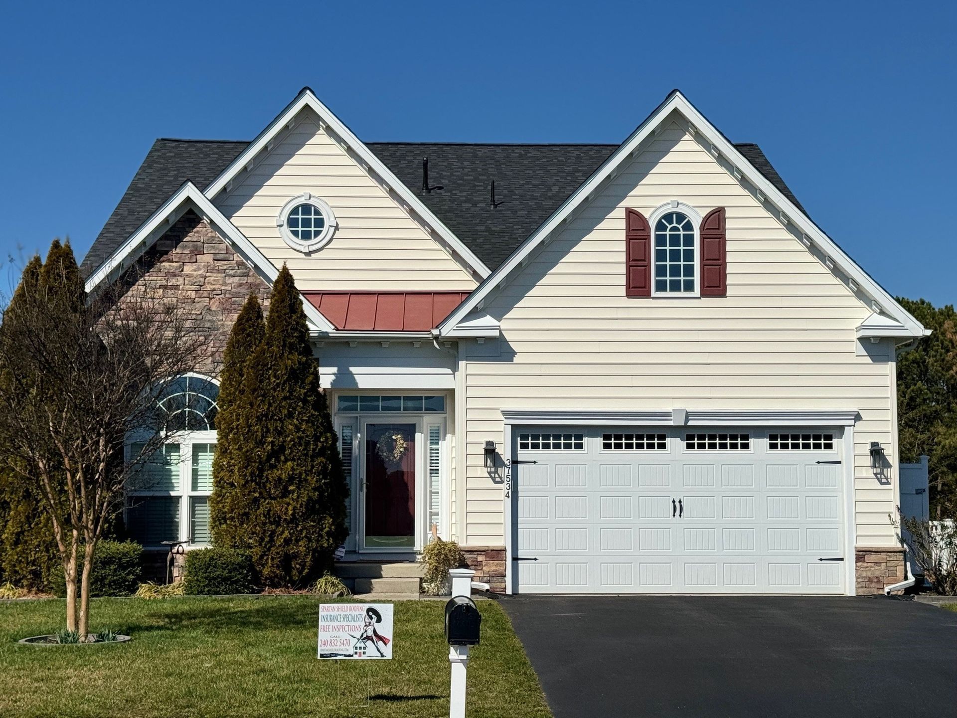 Suburban house with beige siding, white garage, red shutters, and a front lawn under a clear blue sky