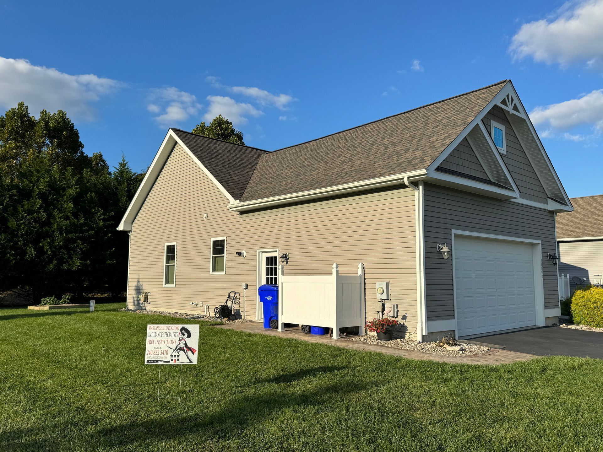 Beige house with gray roof, white garage, blue bin, and lawn sign on a sunny day.