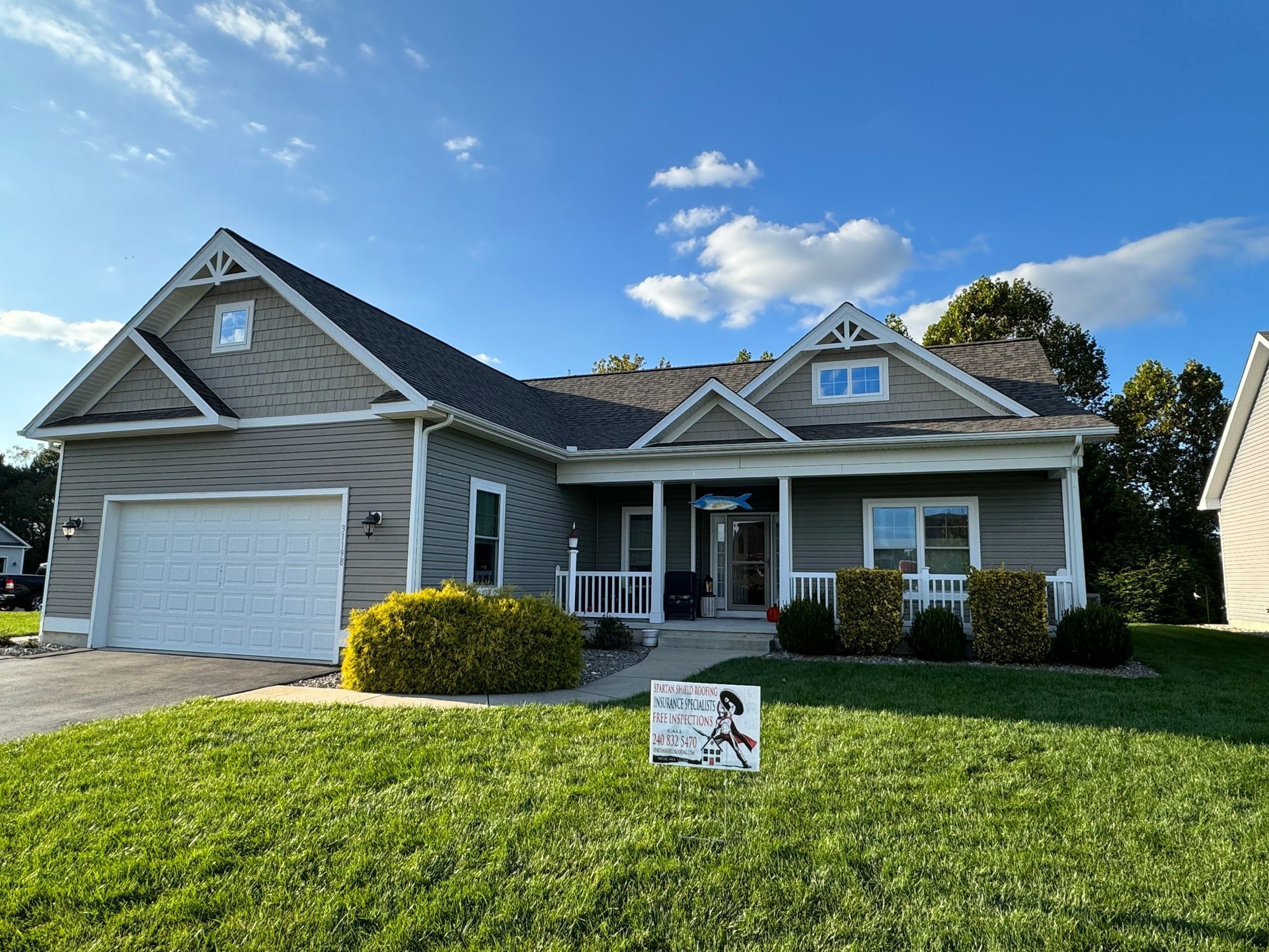Suburban house with gray siding, white garage, front porch, and a real estate sign on the lawn