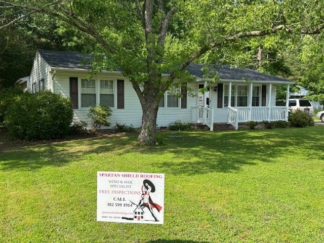 White house with porch and large tree on a sunny lawn, plus a yard sign in front.