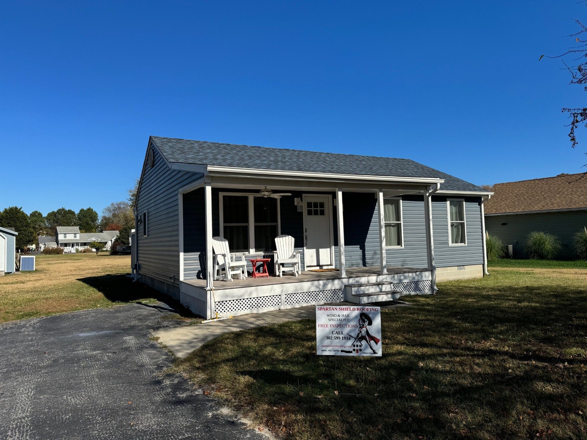 Small gray house with a front porch, white chairs, and a sign on a grassy lot under a blue sky