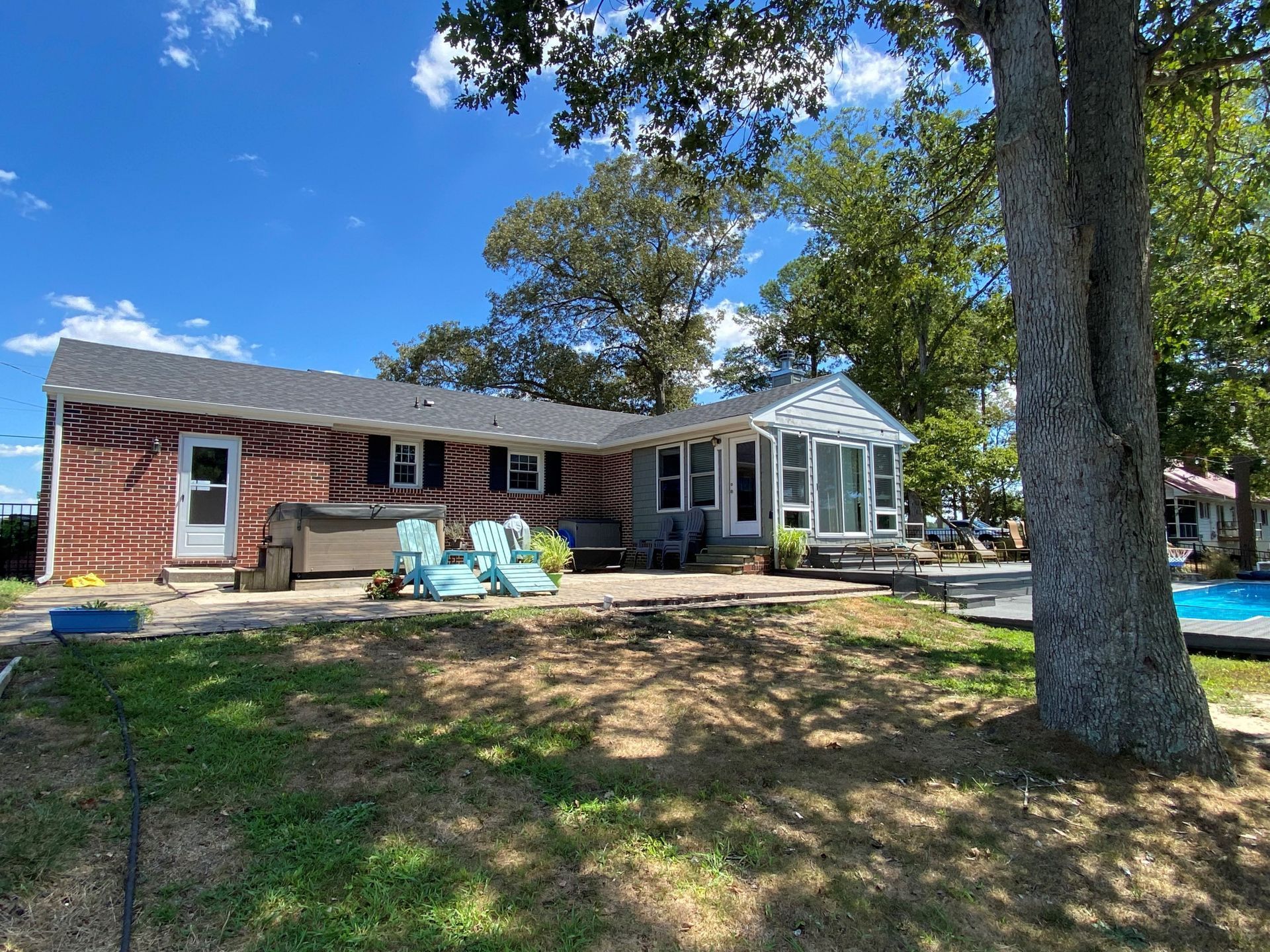 Single-story red brick house with a porch and large tree in the front yard under a blue sky
