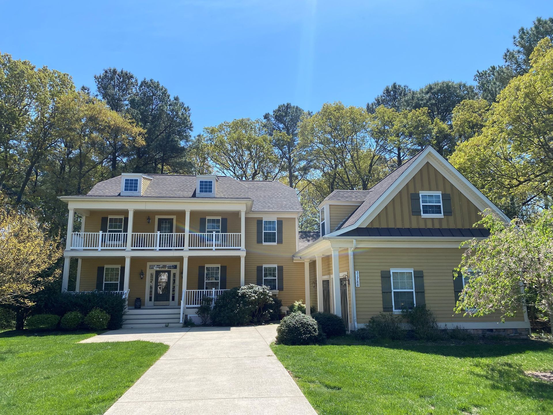 Large beige house with porch and dormers beside a green garage, surrounded by trees under a blue sky