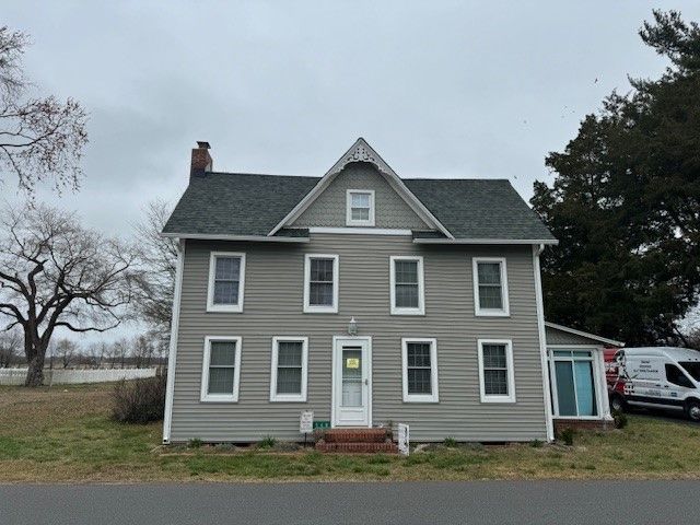 Gray two-story farmhouse with white trim and a steep roof, seen from the front on a cloudy day.