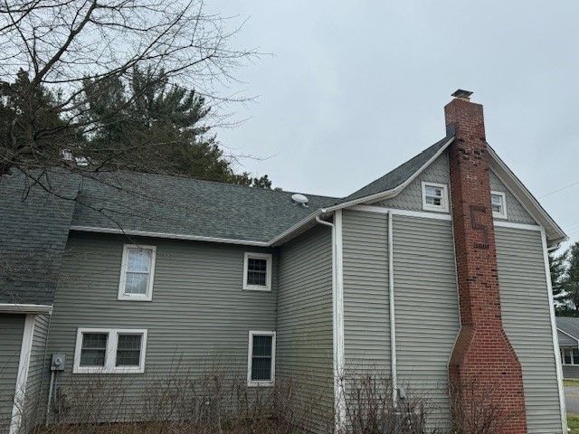 Gray two-story house with a red brick chimney and dark roof in a wooded setting