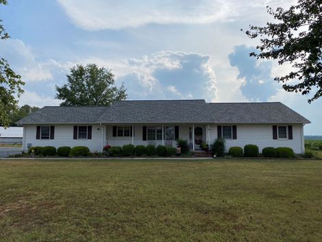 Single-story white house with a gray roof, front porch, and a large grassy yard under a partly cloudy sky