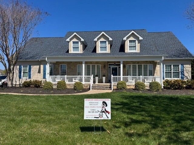 Large blue house with white trim and dormer windows, front porch, and a yard sign on the lawn