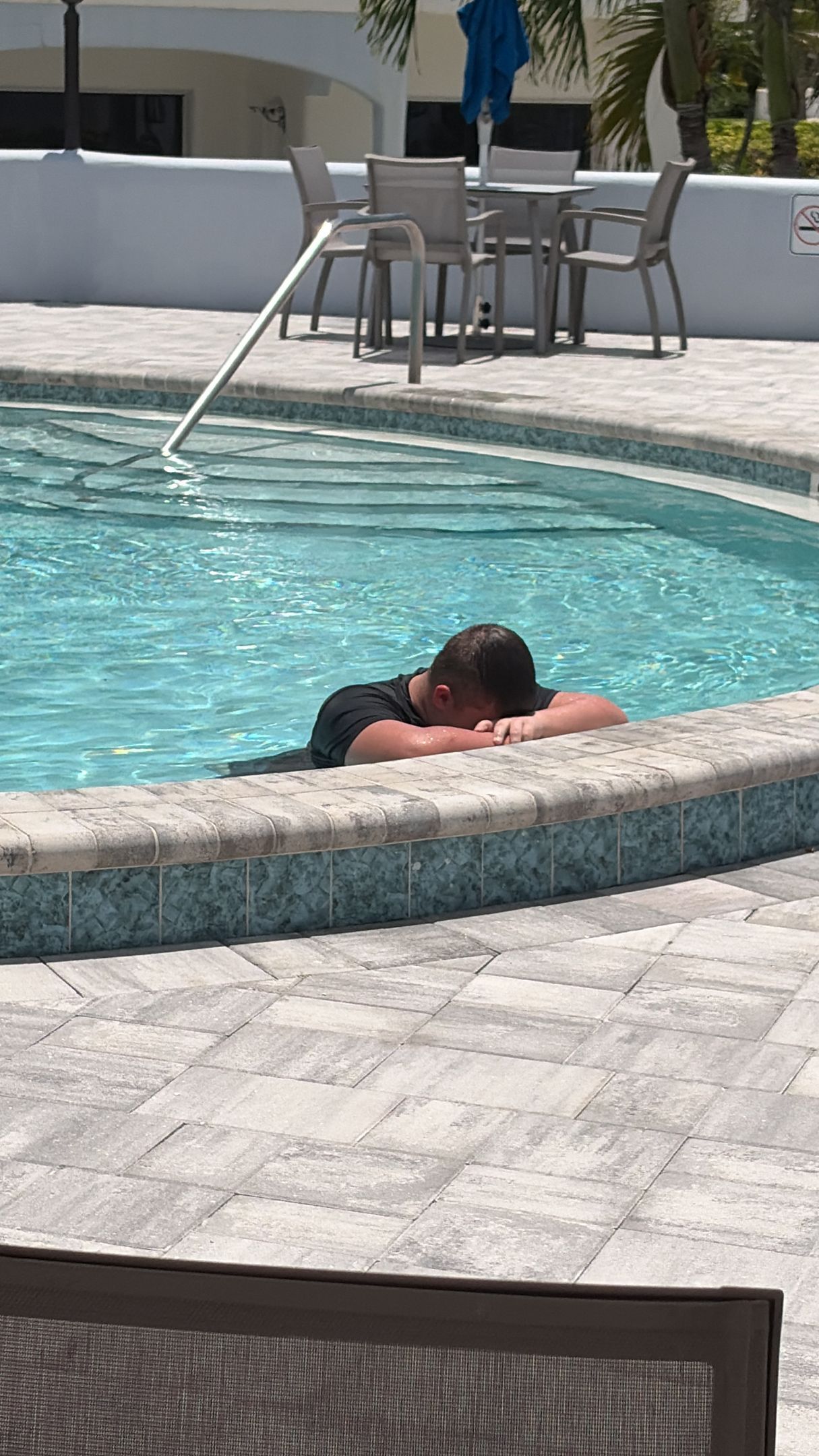 Person resting at the edge of a turquoise swimming pool on a sunny patio.