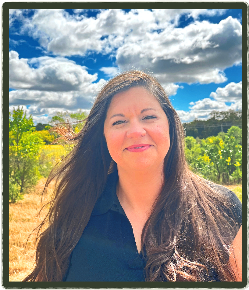 Woman with long brown hair smiling outdoors, blue sky with clouds in the background.