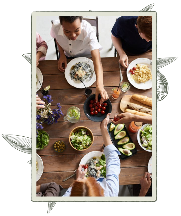 People sharing a meal at a wooden table, passing dishes of food: pasta, salad, avocado, and tomatoes.