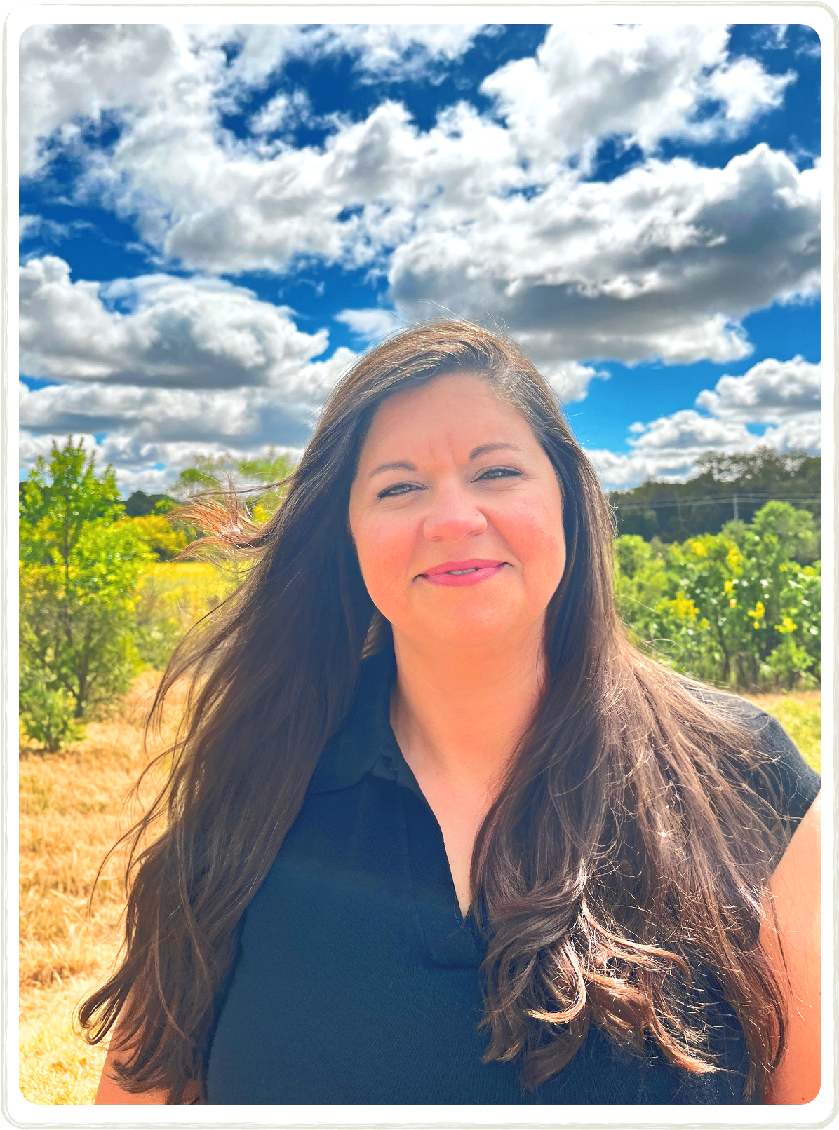 Woman with long brown hair smiles outdoors against a cloudy sky and yellow field.