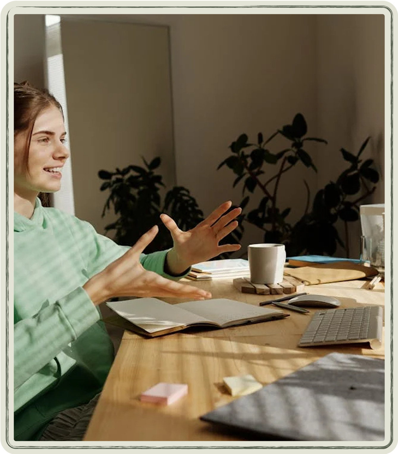 Woman gesturing while speaking, seated at a desk with laptop, notebook, and coffee cup.