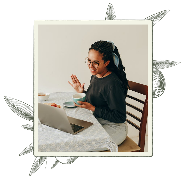 Woman waving at laptop, holding a mug. Seated at table, light-colored clothing, glasses. Interior setting.