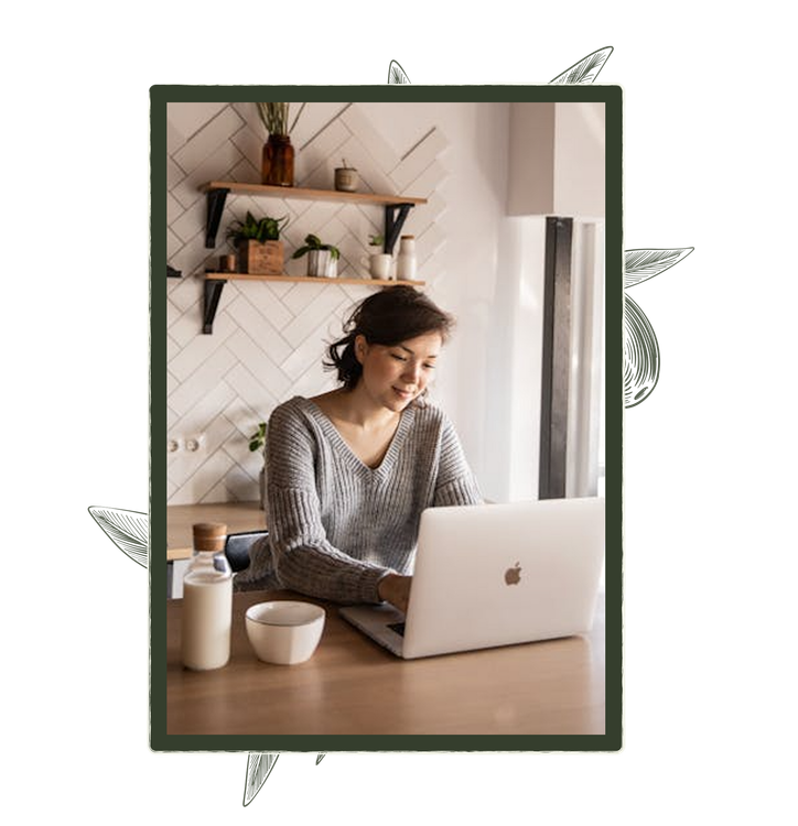 Woman working on a laptop in a kitchen, smiling. A drink and bowl are on the table.