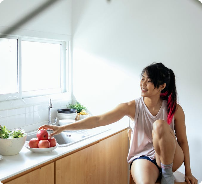 Woman in kitchen reaching for an apple; sitting on counter, smiling, daylight.