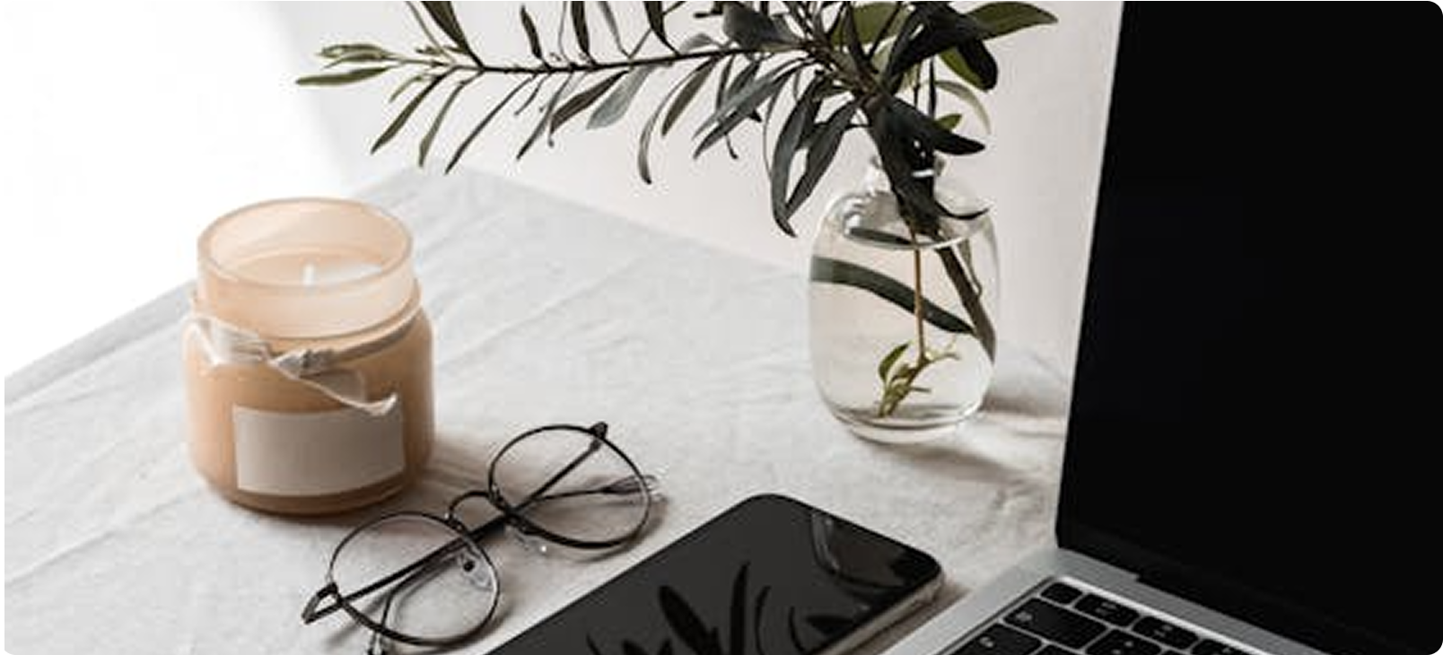 Laptop, smartphone, candle, and glasses on a white surface, with a vase of greenery.