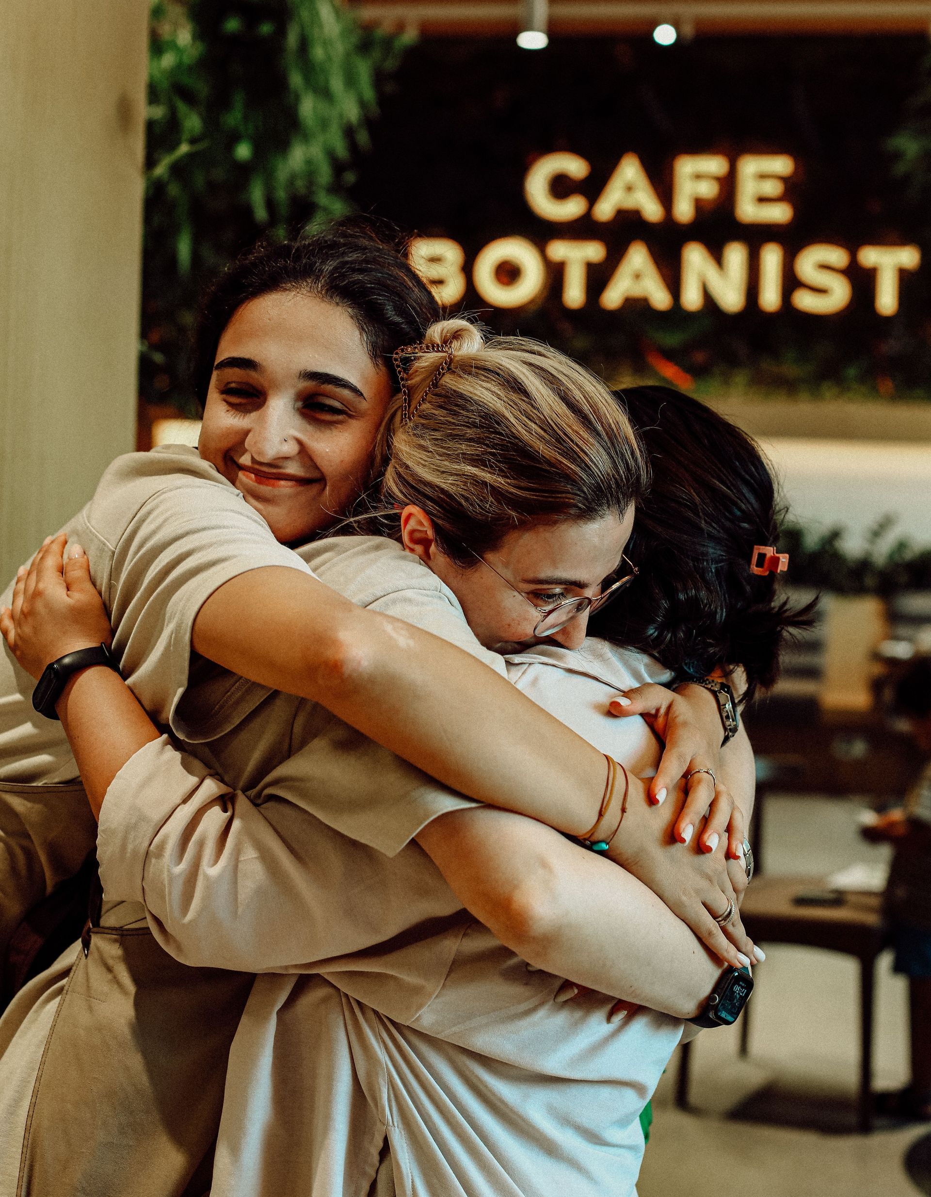 Three friends hugging in front of a café sign reading “Cafe Botanist.”
