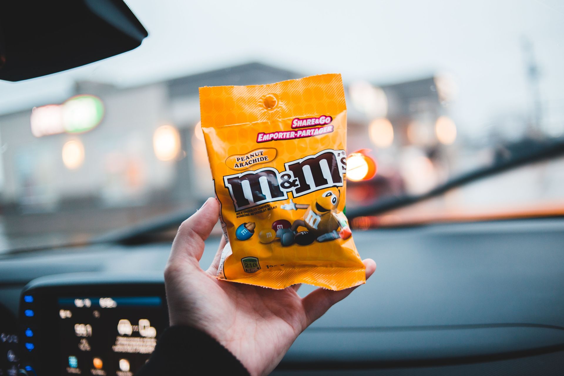 Hand holding a yellow M&M's bag inside a car, with dashboard lights visible.