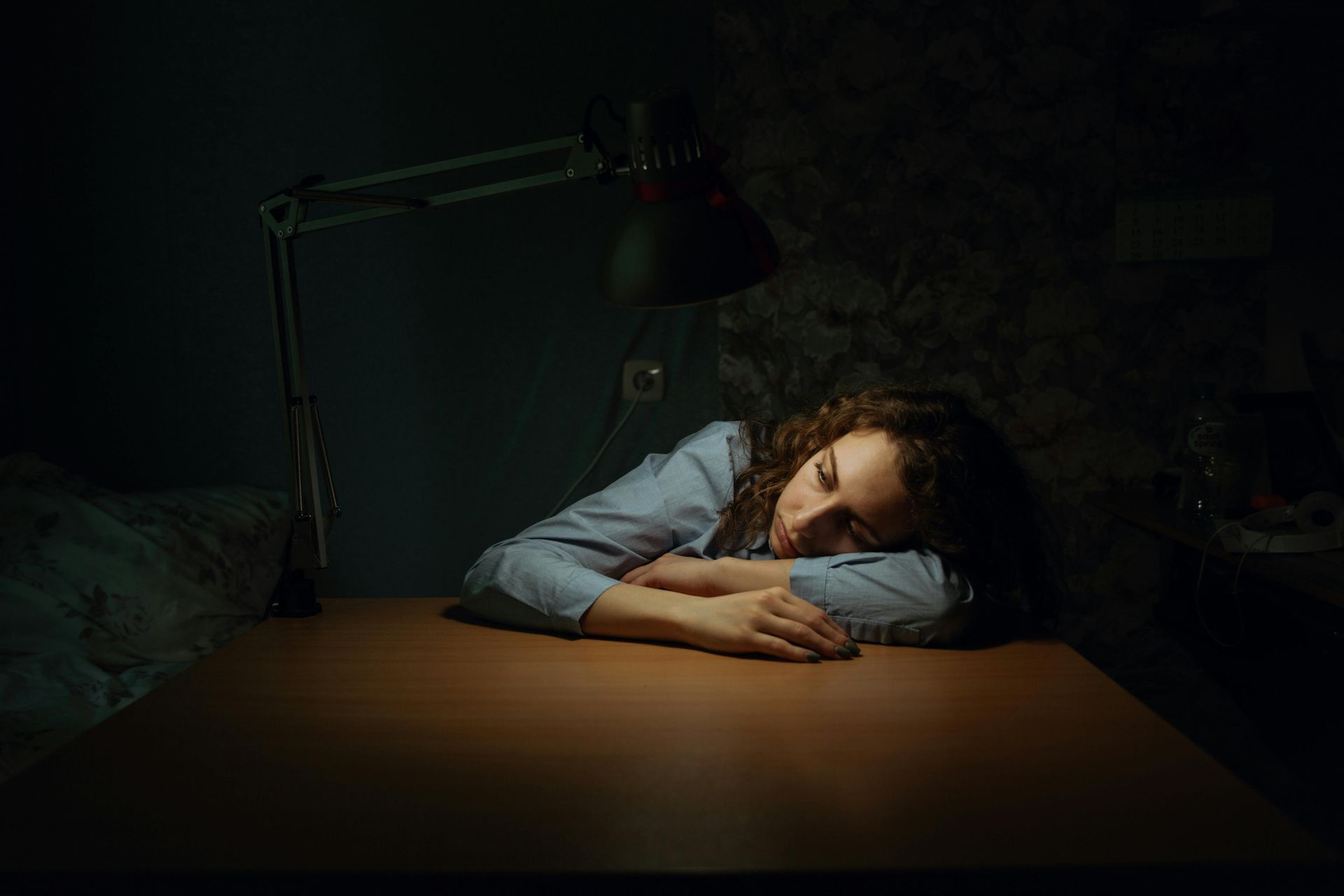 Person asleep at a desk under a dim lamp, resting on folded arms in a dark room