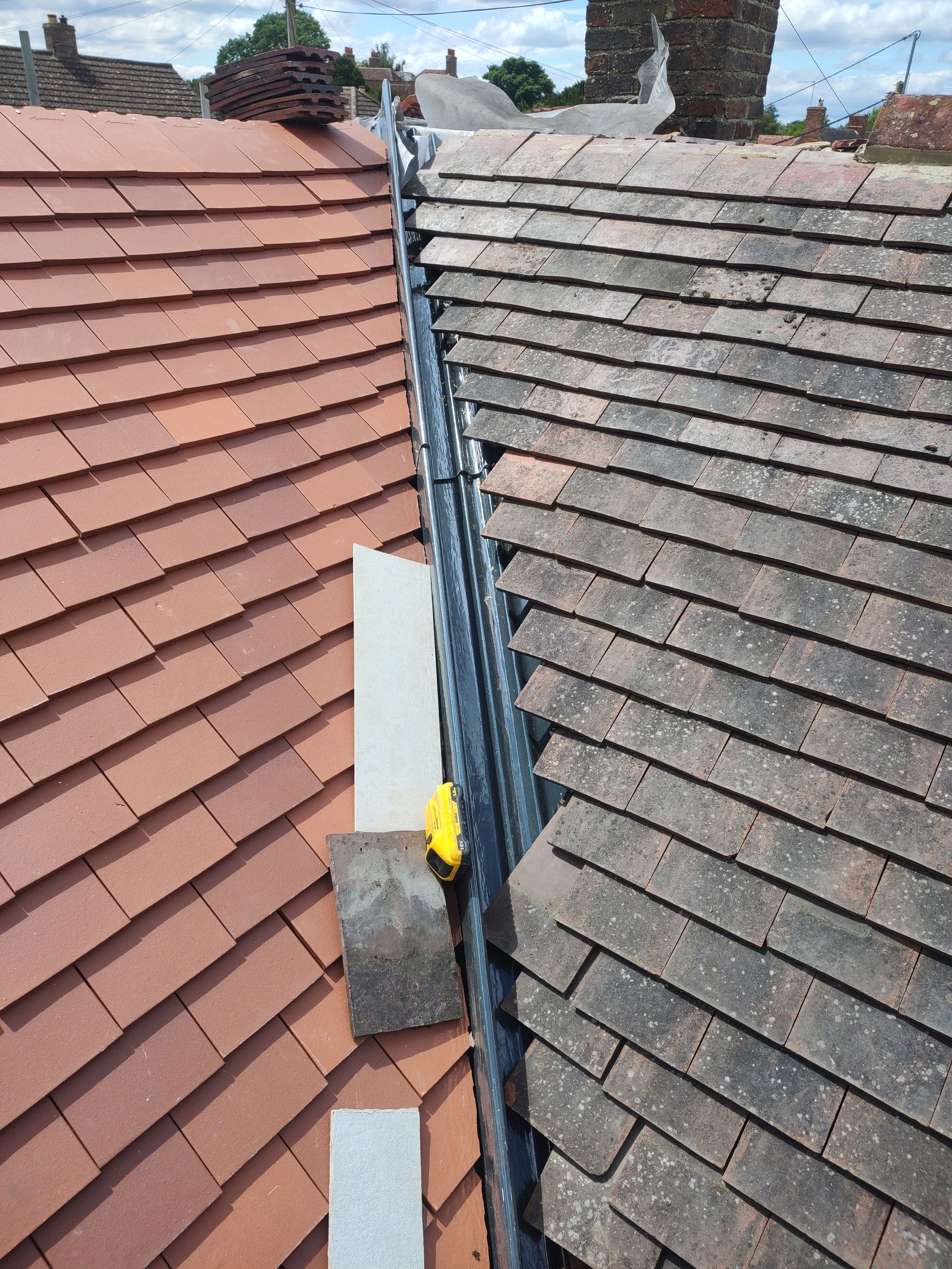 Roof valley between red and gray tiled roofs, with a blue tarp and a yellow tool below.