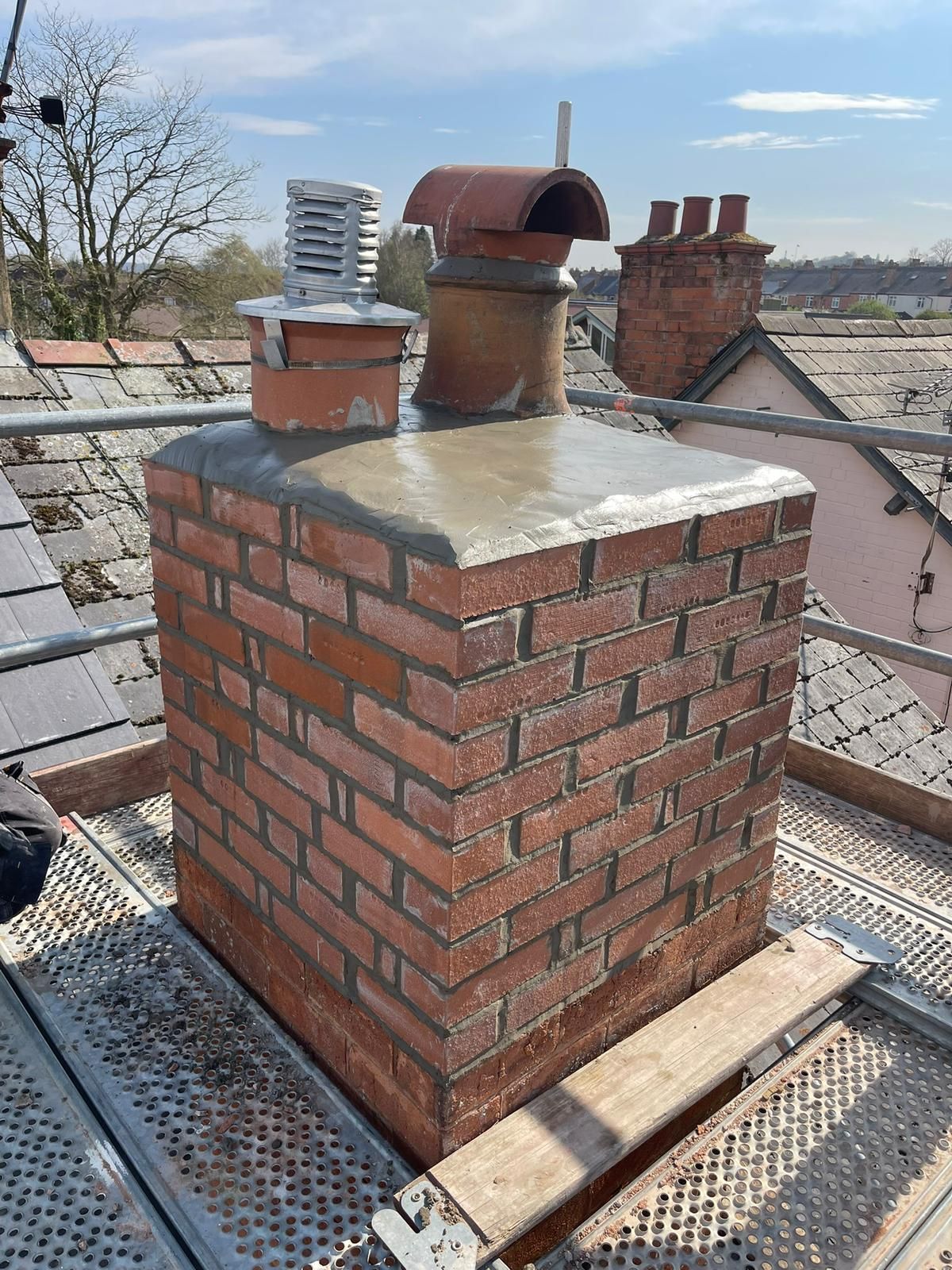 Brick chimney stack on a rooftop with metal cap and surrounding slate roofs under a cloudy sky