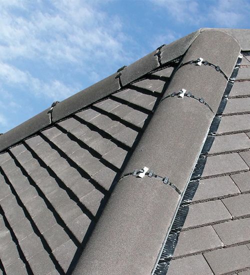 Close-up of a gray shingled roof with a rounded ridge vent and metal roof clips under a blue sky