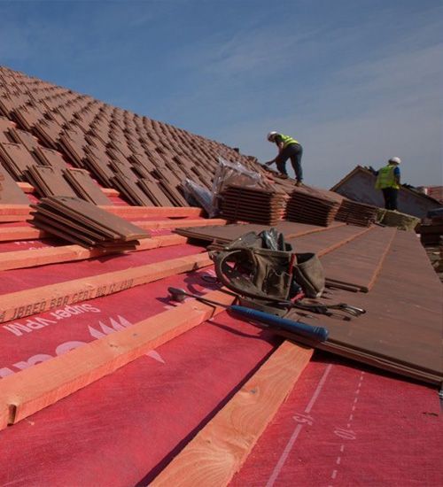 Roofers installing red shingles on a steep roof under a blue sky