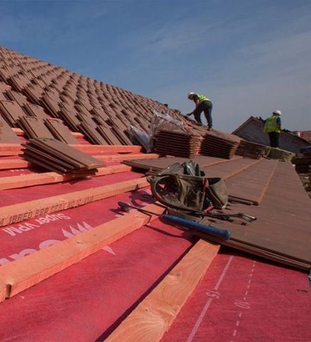 Roofers installing red roofing underlayment and tiles on a steep roof under a clear sky