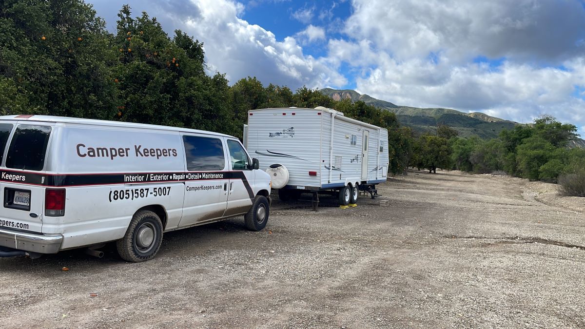 White van towing a travel trailer on a gravel road, with trees and a cloudy sky in the background.
